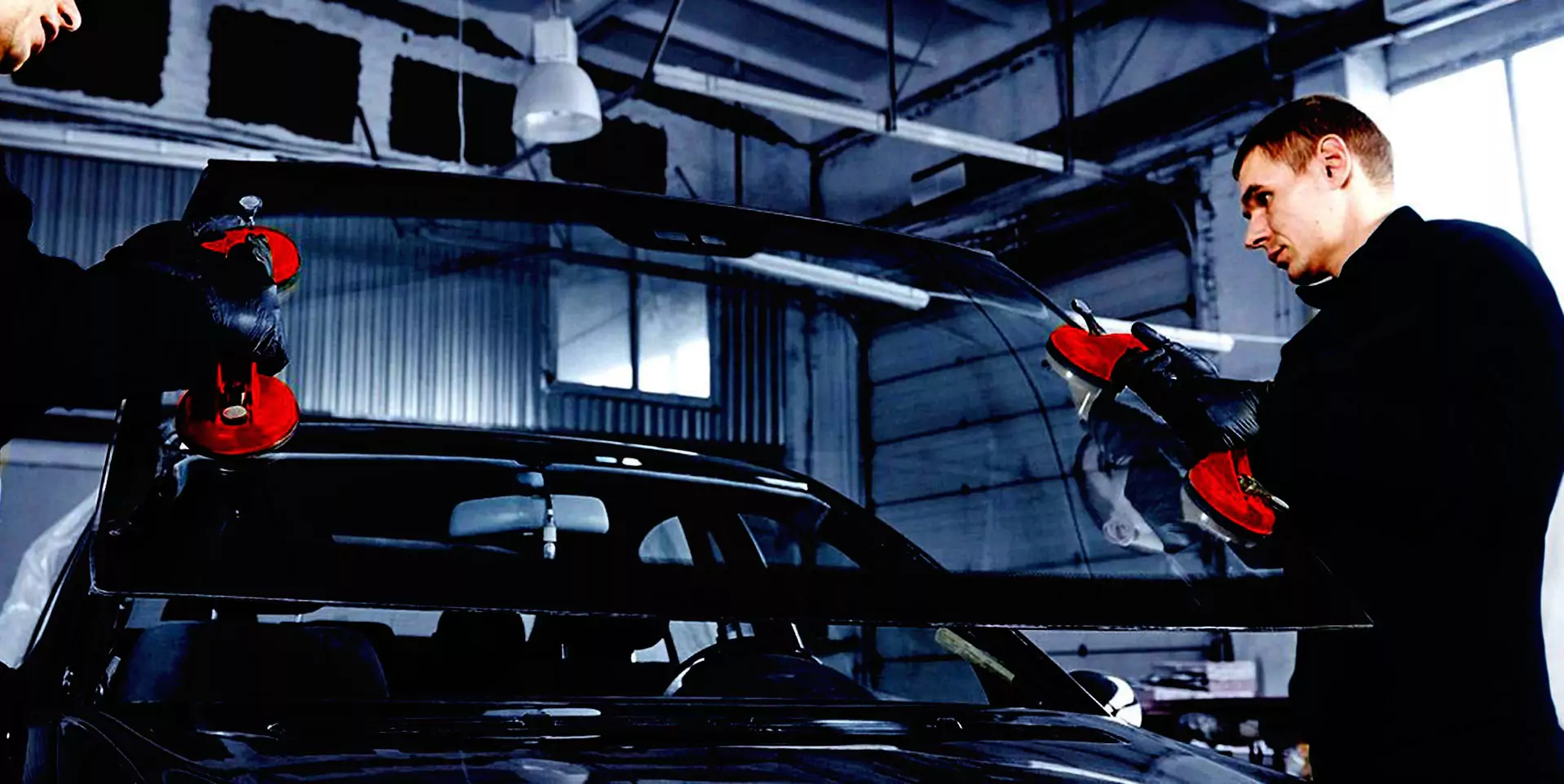 Two technicians install a windshield on a car inside a garage, using suction cups for precise placement