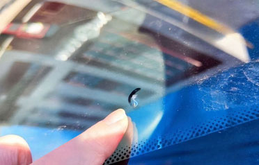 Finger pointing at a small crescent-shaped windshield crack near the edge of the glass